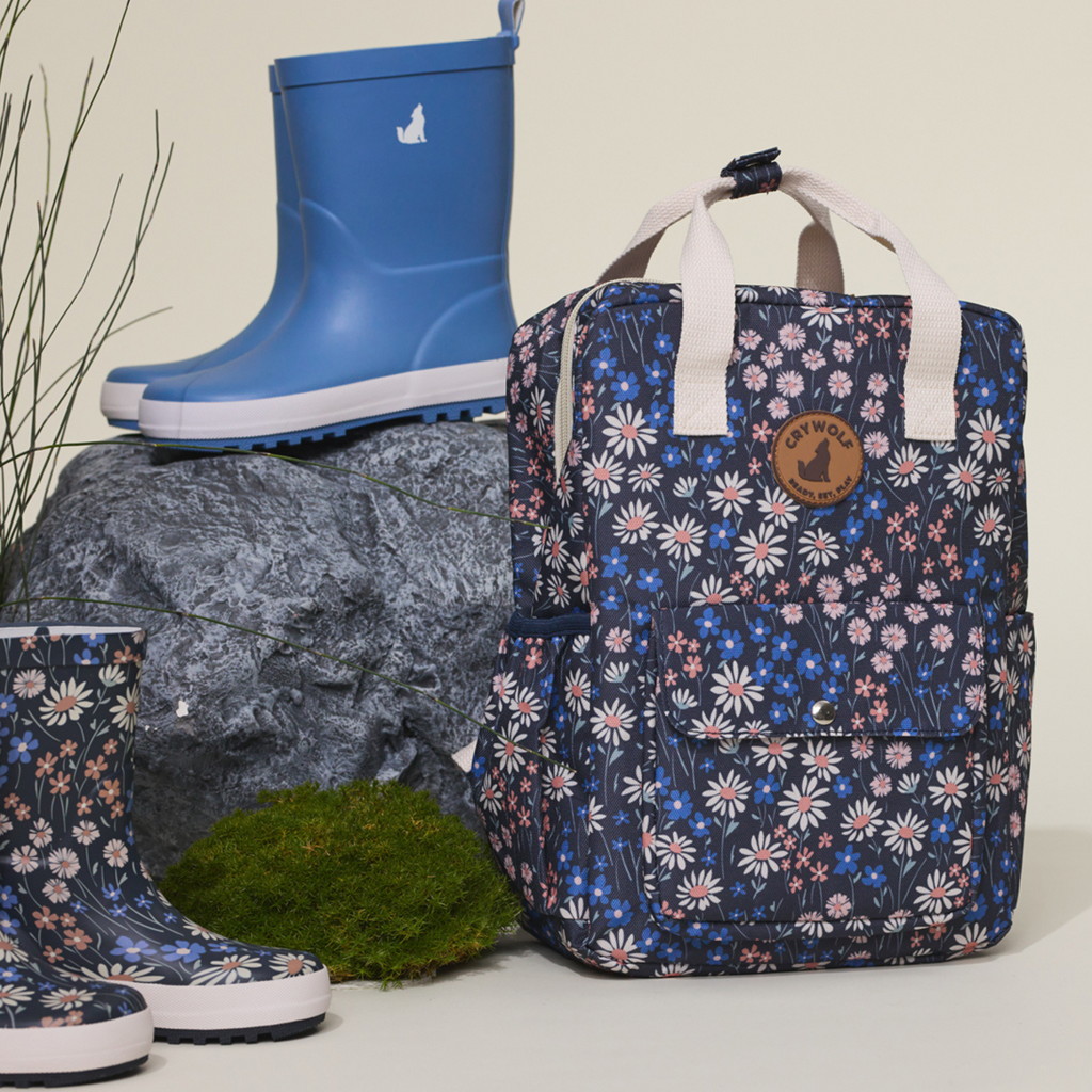 A blue, pink and white floral backpack with matching gumboots and a pair of blue gumboots resting against rocks and foliage indoors