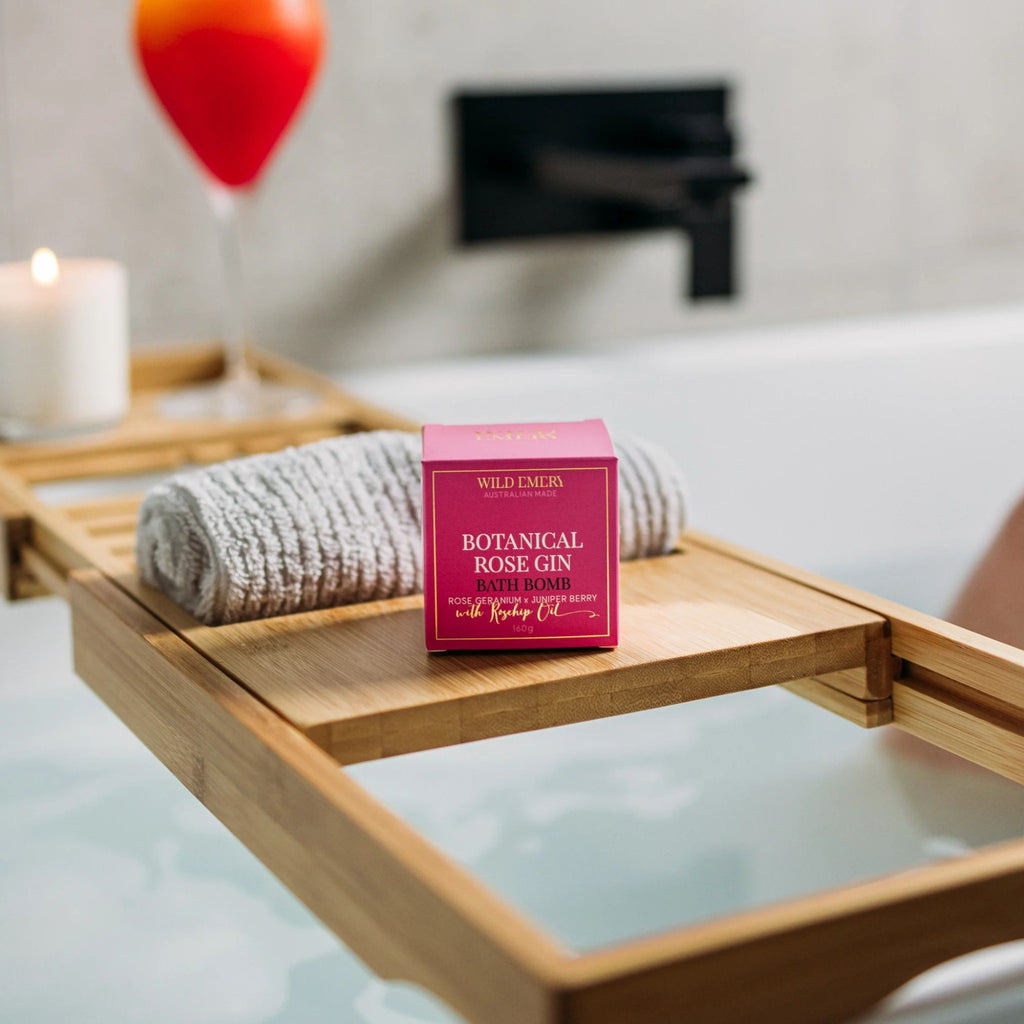 Bath Bomb - Botanical Rose Gin by Wild Emery. Small pink box sitting on a wooden bath shelf over a bathtub. Candle in the background