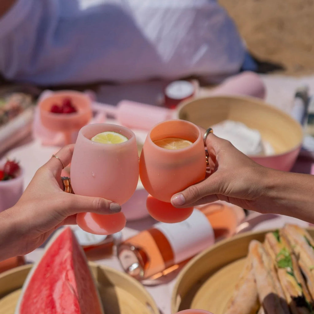 Two hands clinking  a set of Pink and orange silicone wine glasses with a picnic in the background