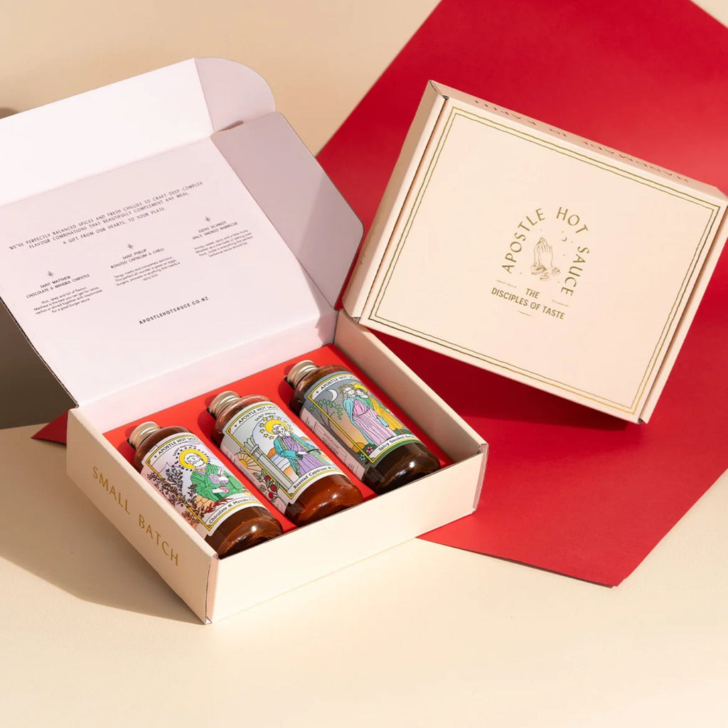 An open box with three bottles of hot sauce in it and a closed box labeled "apostle hot sauce The Disciples of Taste" on a pink and red background