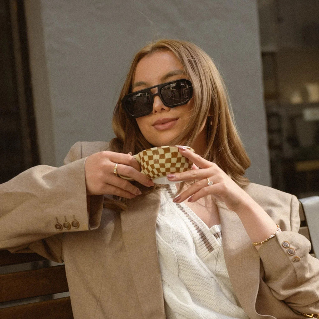 A woman holding a mug outside wearing a blazer vest and sunglasses