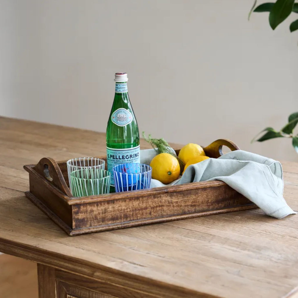 Wood tray of three striped tumbler glasses in varied colours with lemons, a tea towel and bottle of water