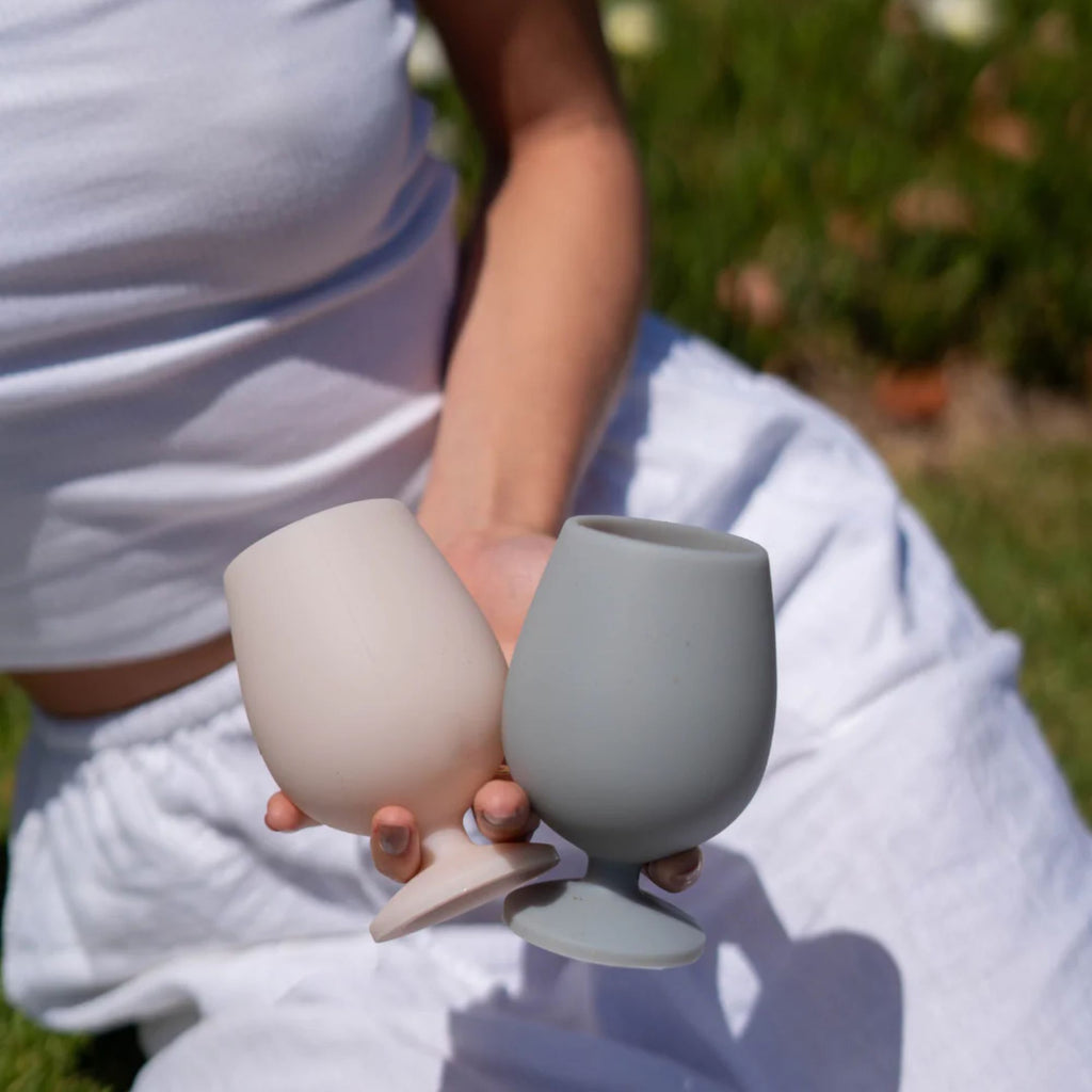 A woman with her head out of frame in white sitting outside holding a set of two short silicone wine glasses in the colours cream and grey in one hand