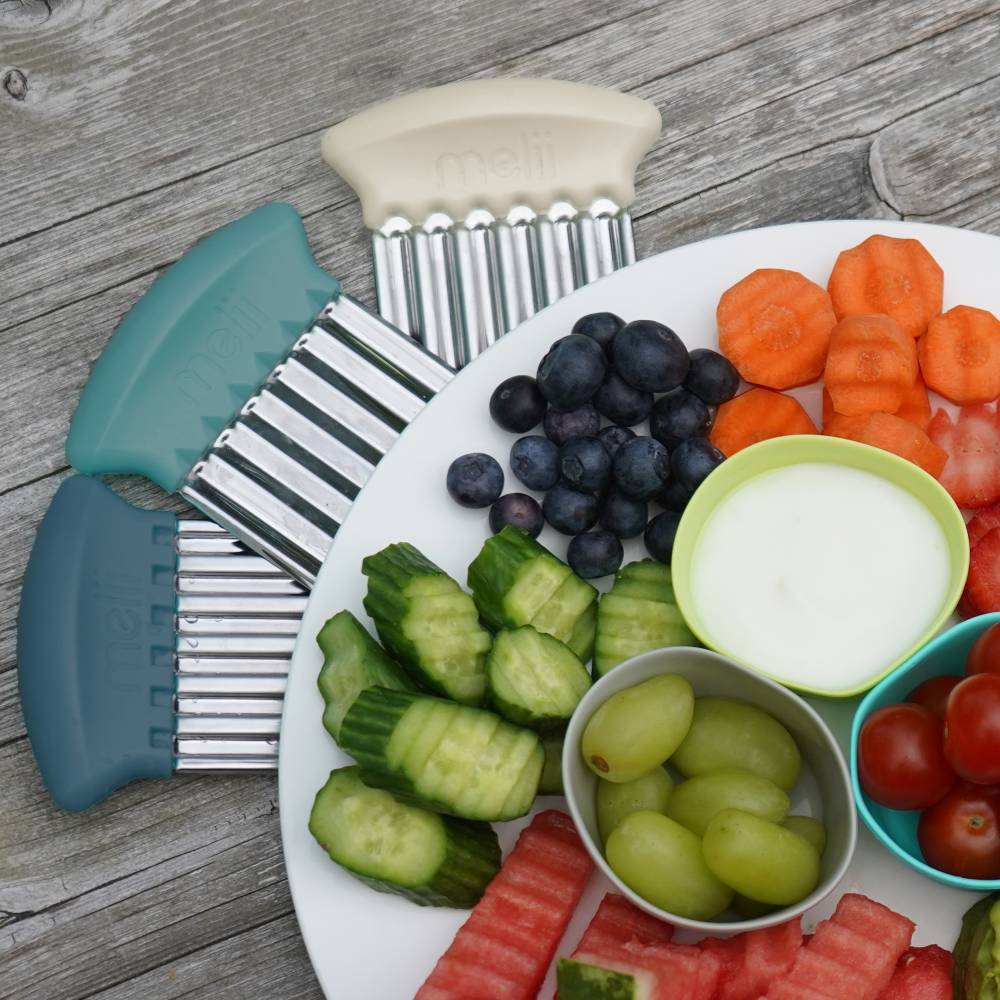 Hand held food cutters with light blue, white and dark blue handles next to a plate of fruit and vegetable snacks