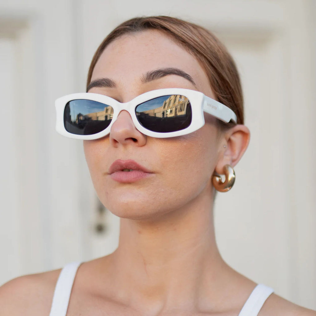 A woman wearing Bored George Sydney white sunglasses with a solid white frame and dark lens and gold hoop earrings