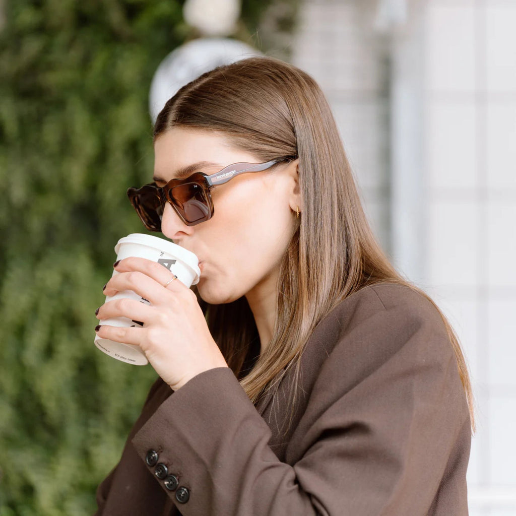 A woman in a brown blazer ad sunglasses sipping a takeaway coffee cup 