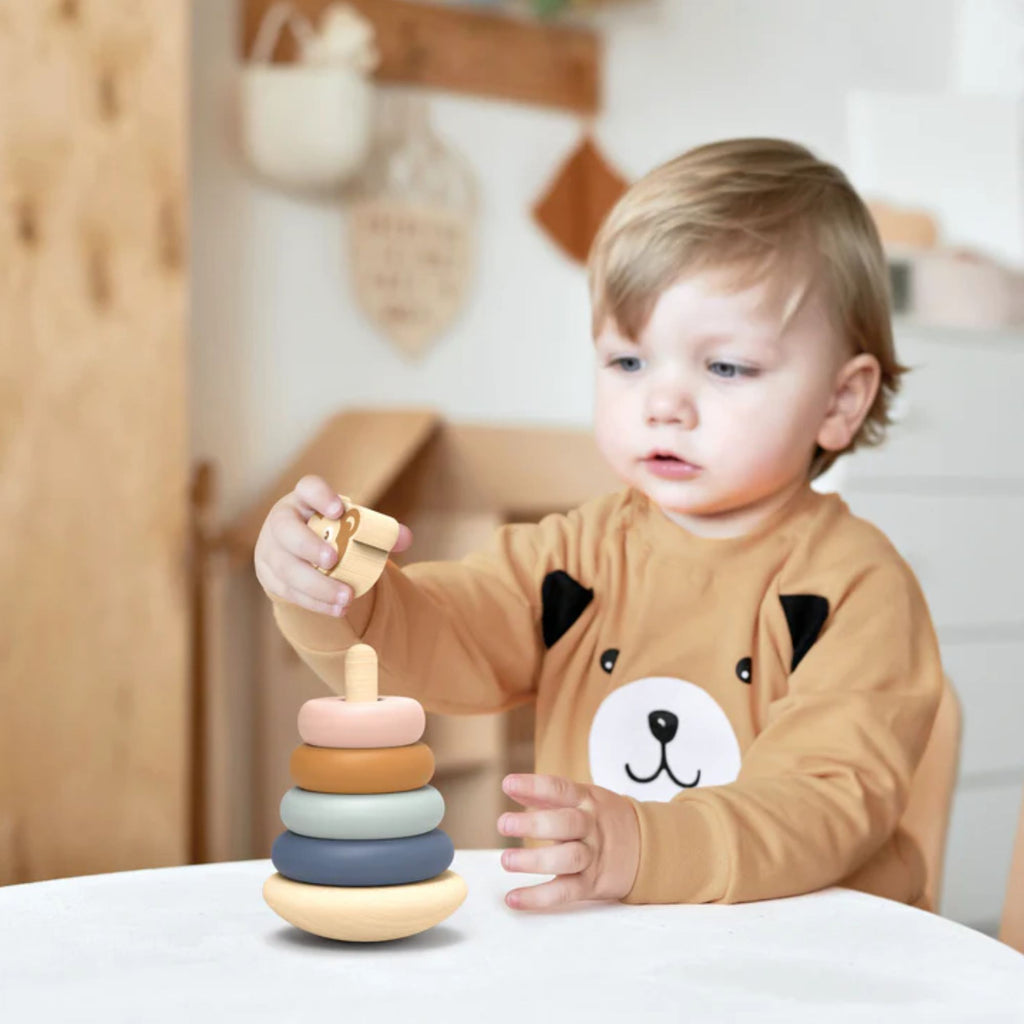 A young boy laying indoors with a set of wooden stacking ring toys in different colours with a monkey on the top 