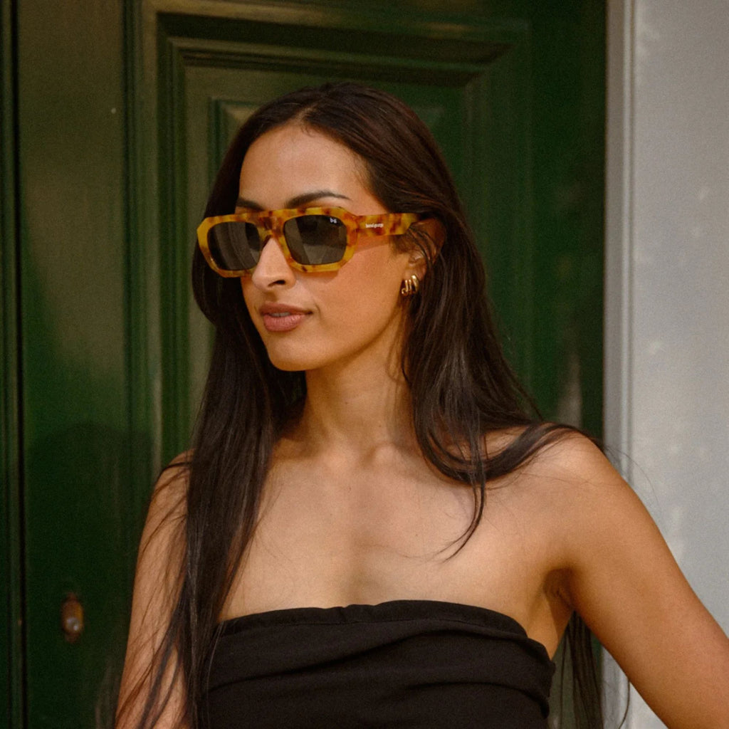 A woman in a blank tank top wearing Bored George Ned orange sunglasses with an orange tort frame and dark lens resting against a green door