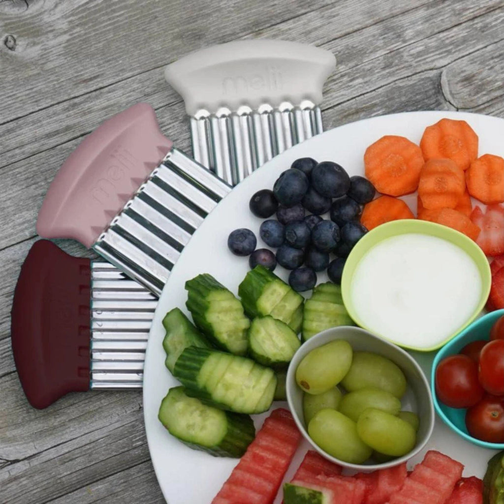 Hand held food cutters with light pink, white and dark red handles  next to a plate of fruit and vegetable snacks