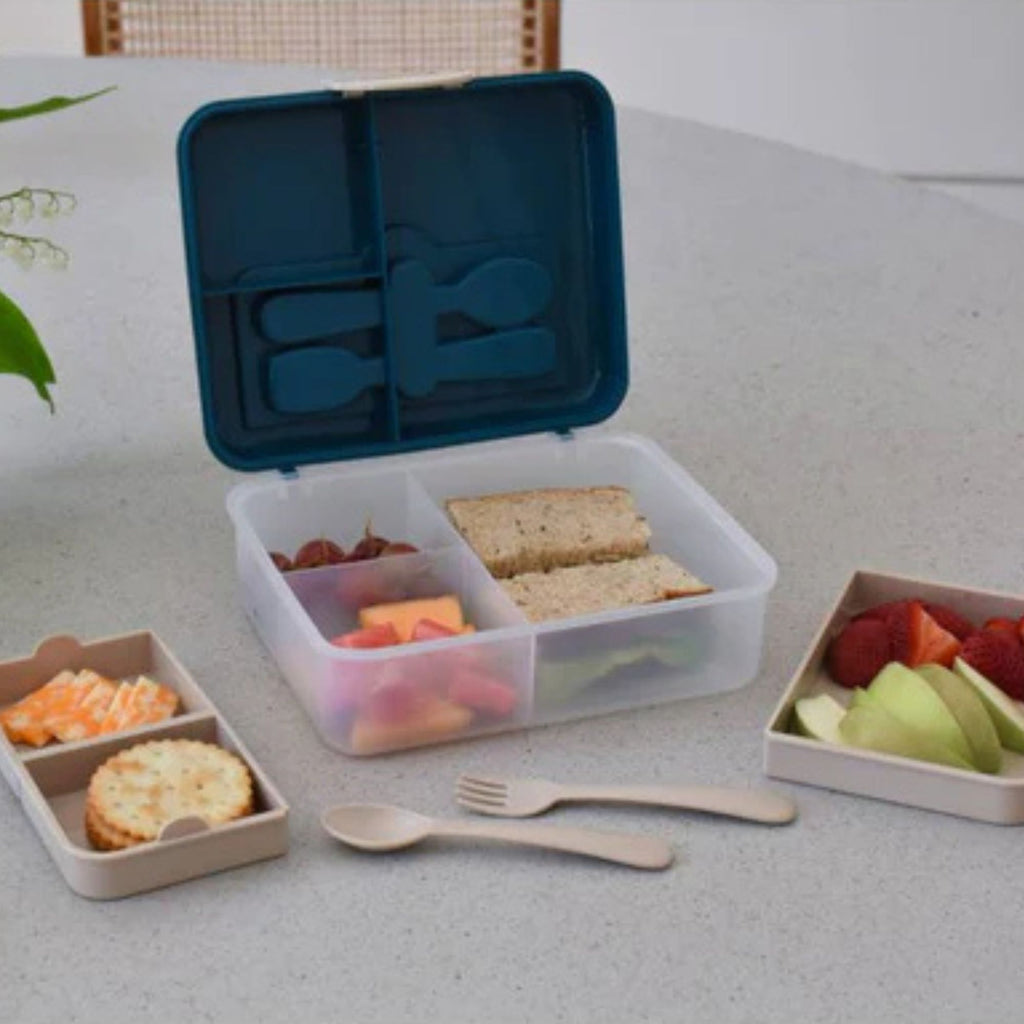 An open plastic lunchbox with a dark blue lid and transparent bottom filled with snacks featuring removable compartments and cutlery 