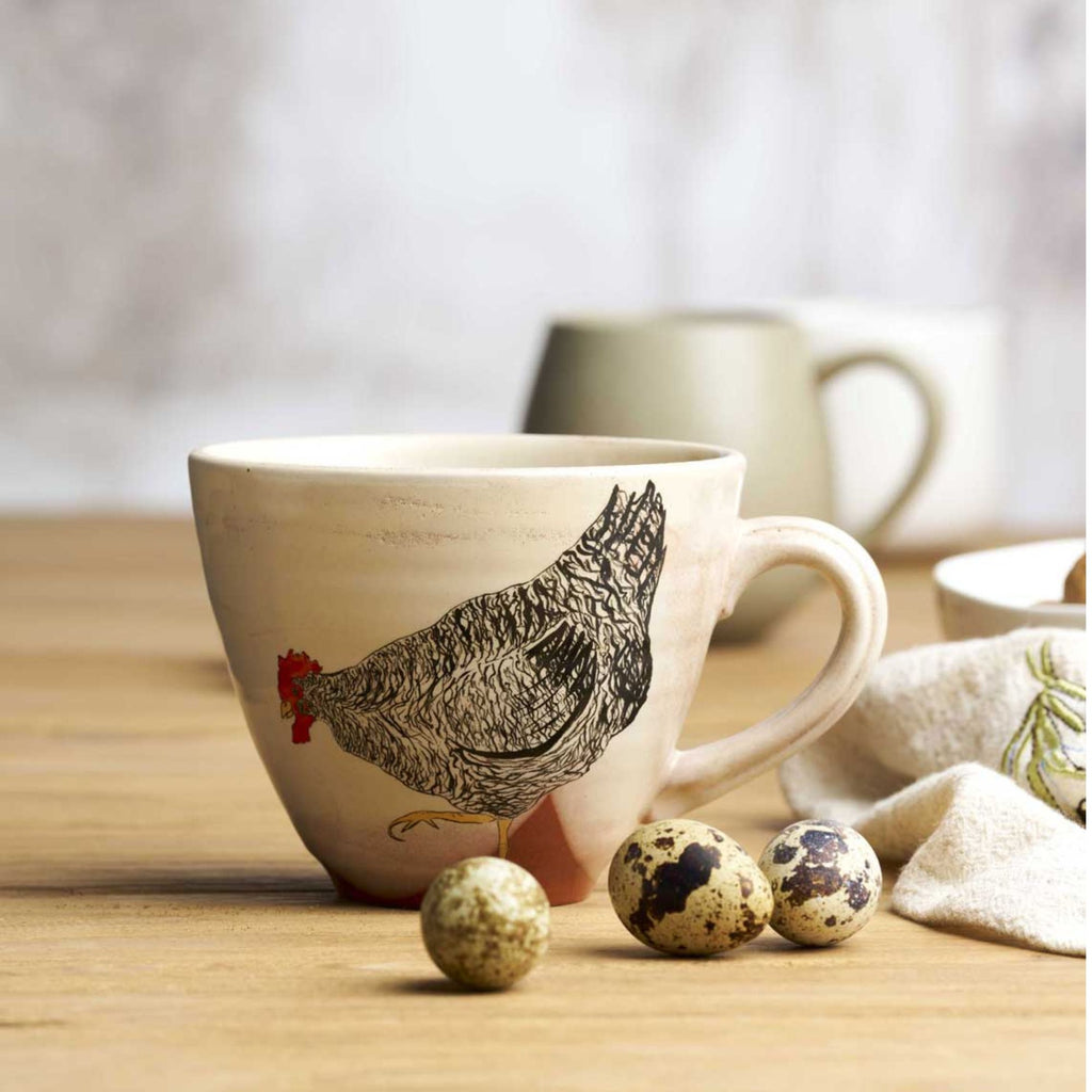 Wide white mug with a chicken print, some eggs, a tea towel and light grey mug 