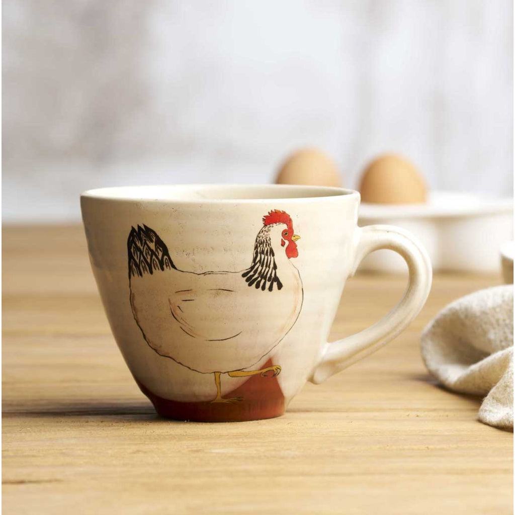 A white white mug with a chicken illustration printed on it, eggs and a tea towel