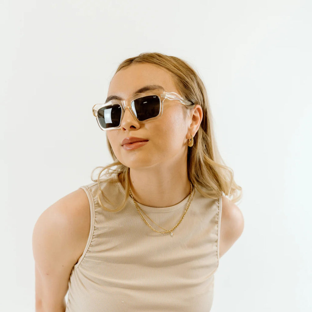 A woman wearing sunglasses, gold necklaces and a beige tank top looking at the camera