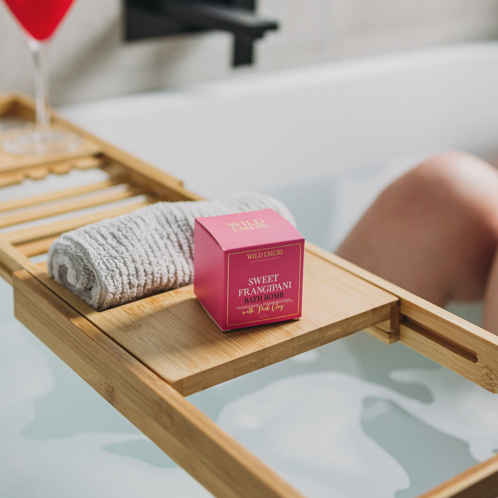 Bath Bomb - Sweet Frangipani by Wild Emery. A pink box on a wooden tray shelf over a bathrub