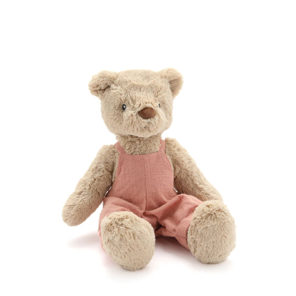 A beige teddy bear in pink linen overalls sitting against a white background