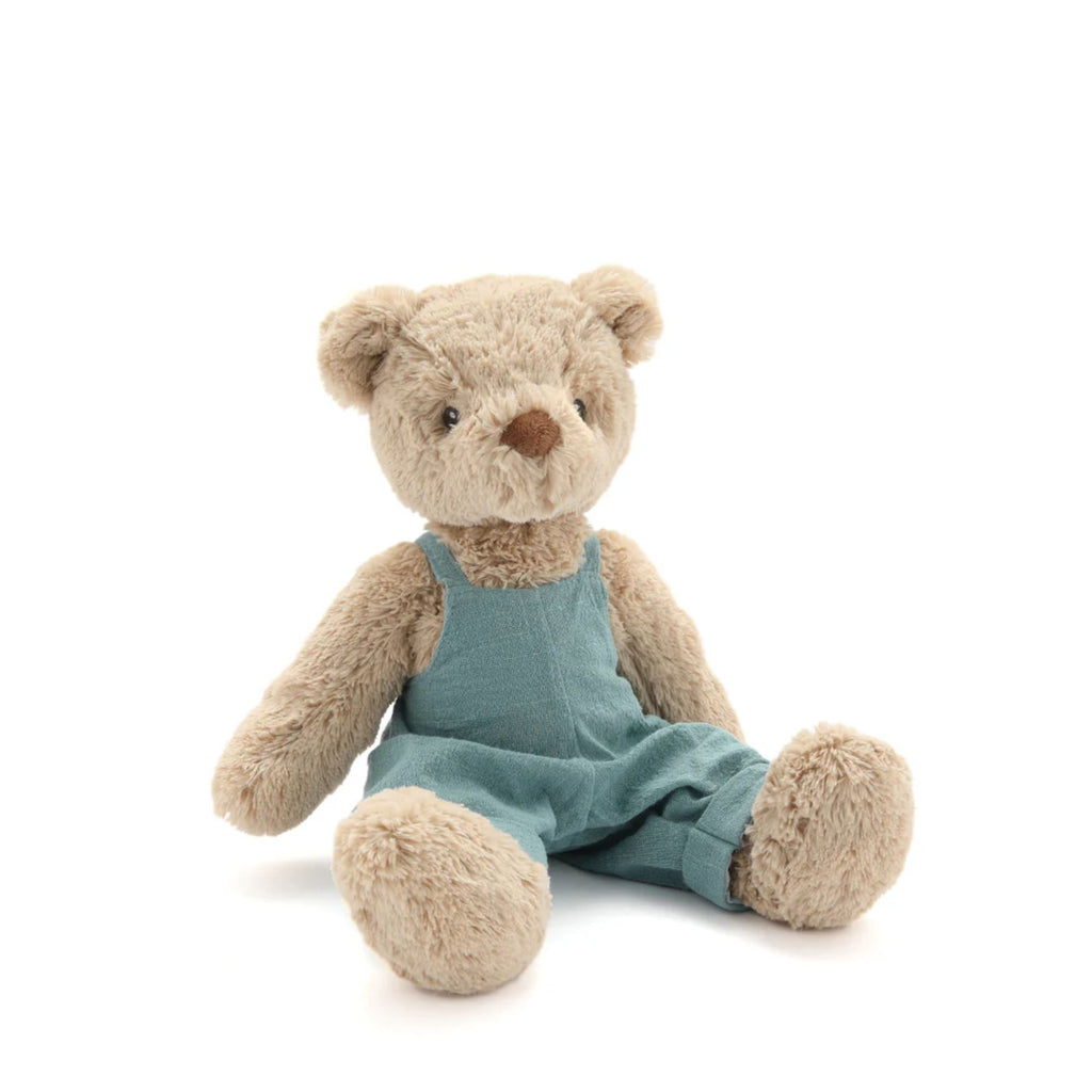 A beige teddy bear in blue linen overalls sitting against a white background