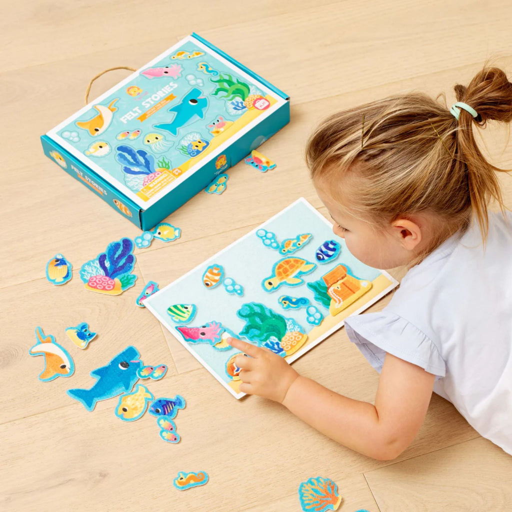 A Cardboard box set placed on the floor with illustrations of ocean creatures labelled "felt stories under the sea" and a girl lying on the floor a felt underwater setting board with felt fish