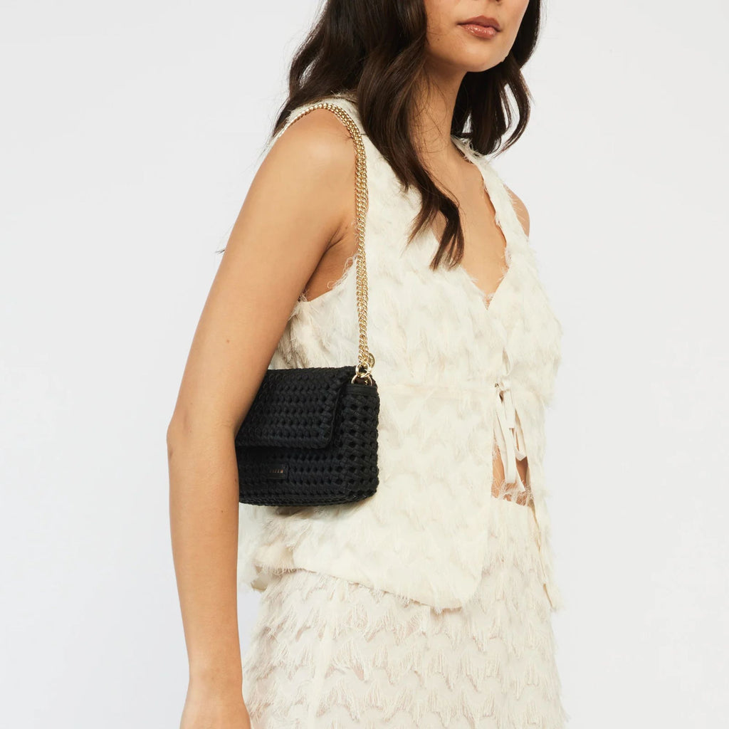 A woman in an all white matching fringe tie up outfit with a black Saben brand baguette shaped purse composed of braided black leather with a gold chain on her shoulder