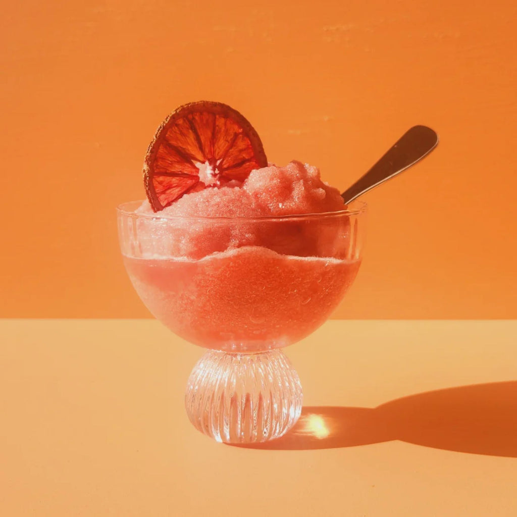 A orange slushy in a clear glass with a dried citrus slice and spoon against an orange background 