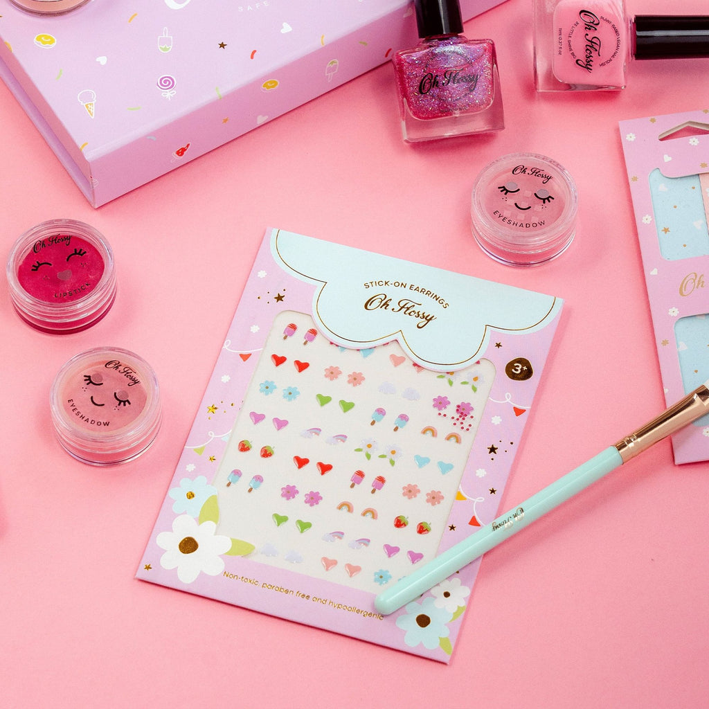 A sheet if various children's sicker earrings visible through a transparent sheet surrounded by pink packaging with flowered labelled "oh Flossy" with the subtitle "stick on earrings" surrounded by various cosmetics including an eyeshadow brush, eyeshadow pots in pink and pink nail polish on a pink table 