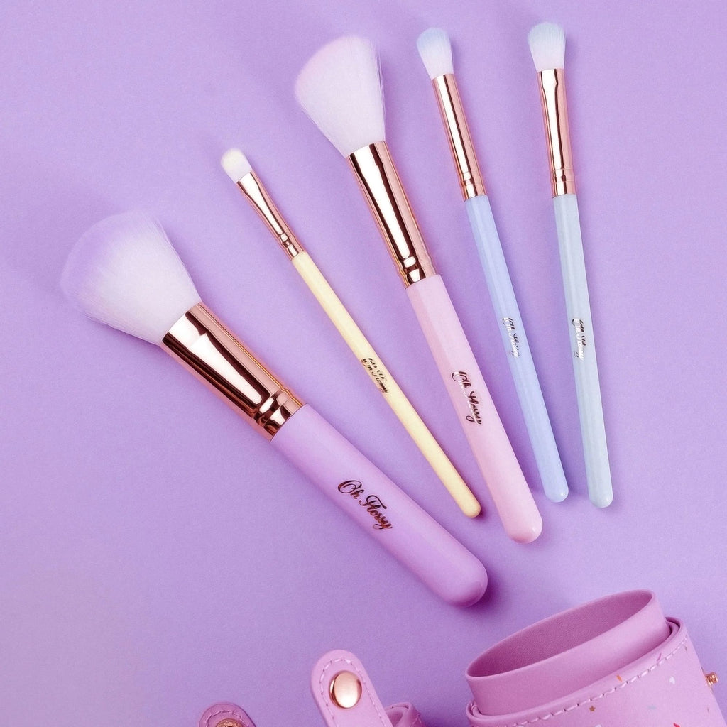 A purple leather cylinder case and 5 various makeup brushes printed with Oh Flossy gold branding lying against a purple background 