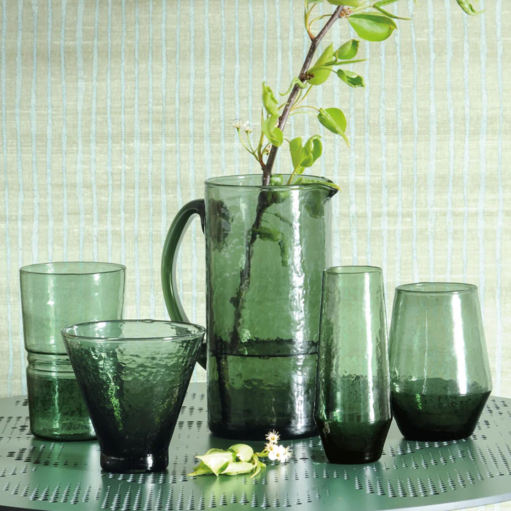 A set of green hammered texture glassware featuring a highball tumbler, drinking tumbler, a jug with a stem and water in it, a stemless champagne flute and a stemless wine glass on a table with a textured wallpaper background