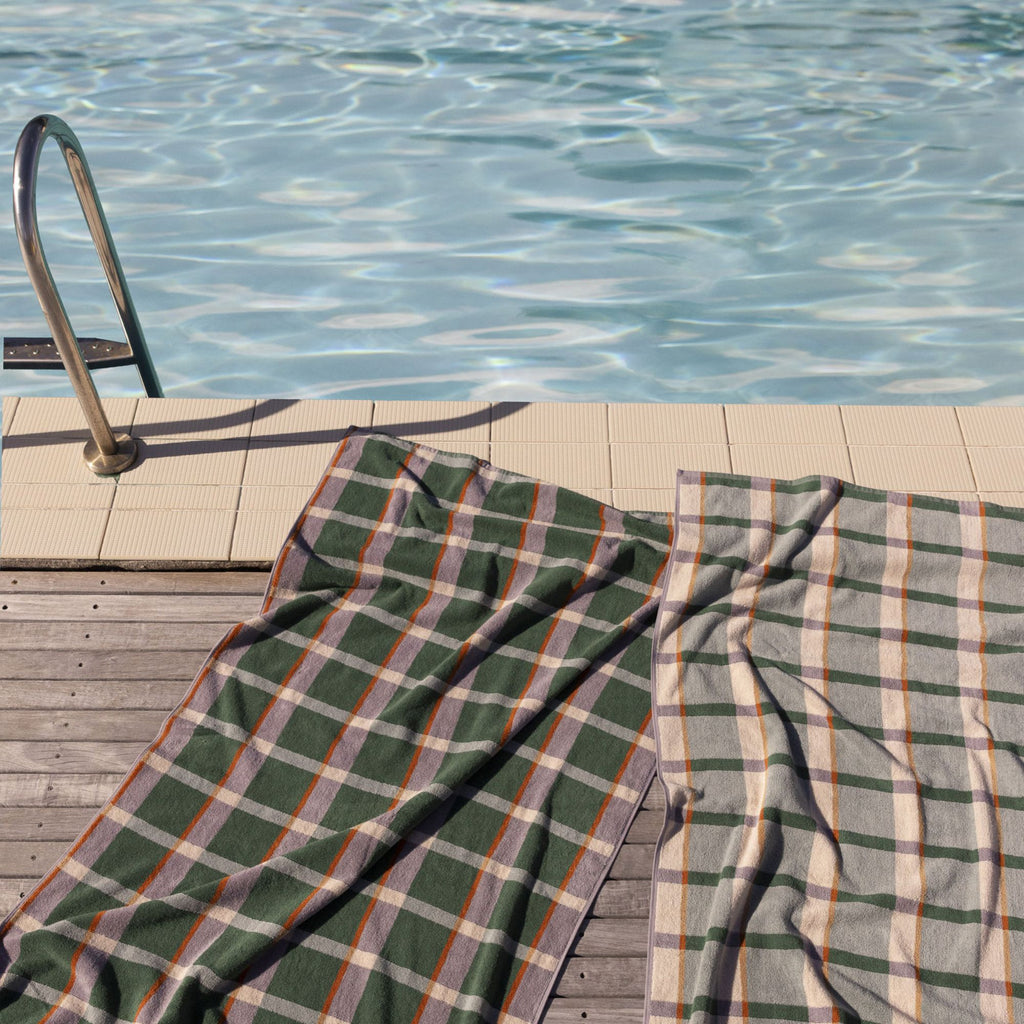 Two multicoloured check towels resting on a wooden deck and white tile bordered pool with a ladder into it 