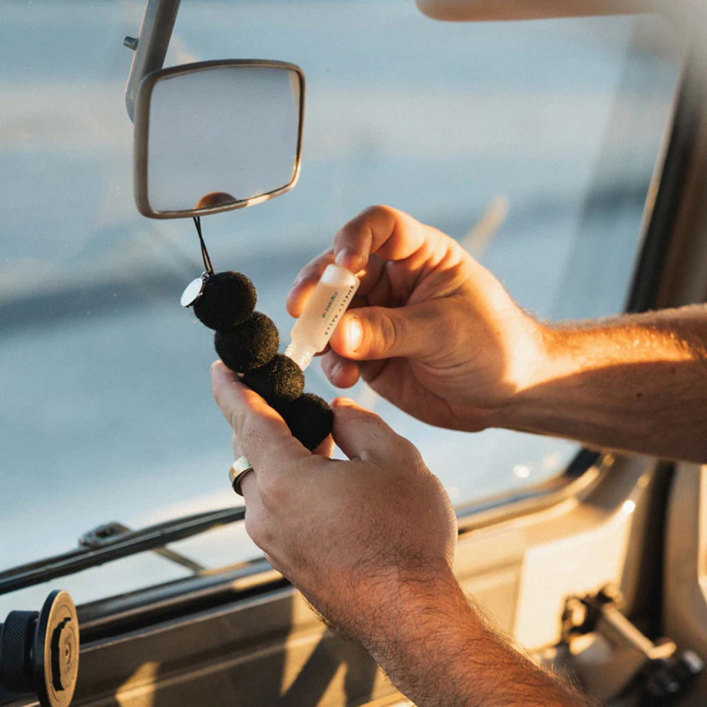 Hands in a car refilling a felt ball car air freshener hanging from a rearview mirror with fragrance oil