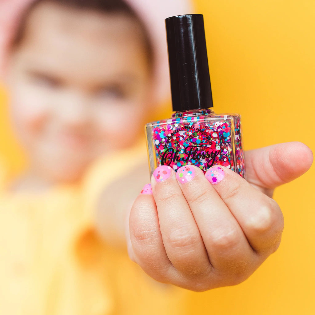 A picture of a blurred girl with a focused hand holding a bottle of rainbow glitter nail polish with painted nails against a yellow background 