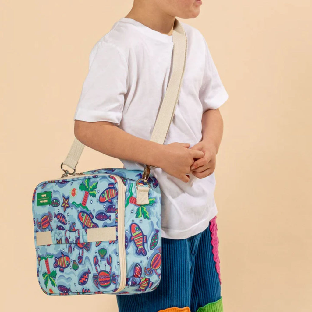 Boy wearing a blue lunch box with painted sea creatures and a cream strap slung over his body 