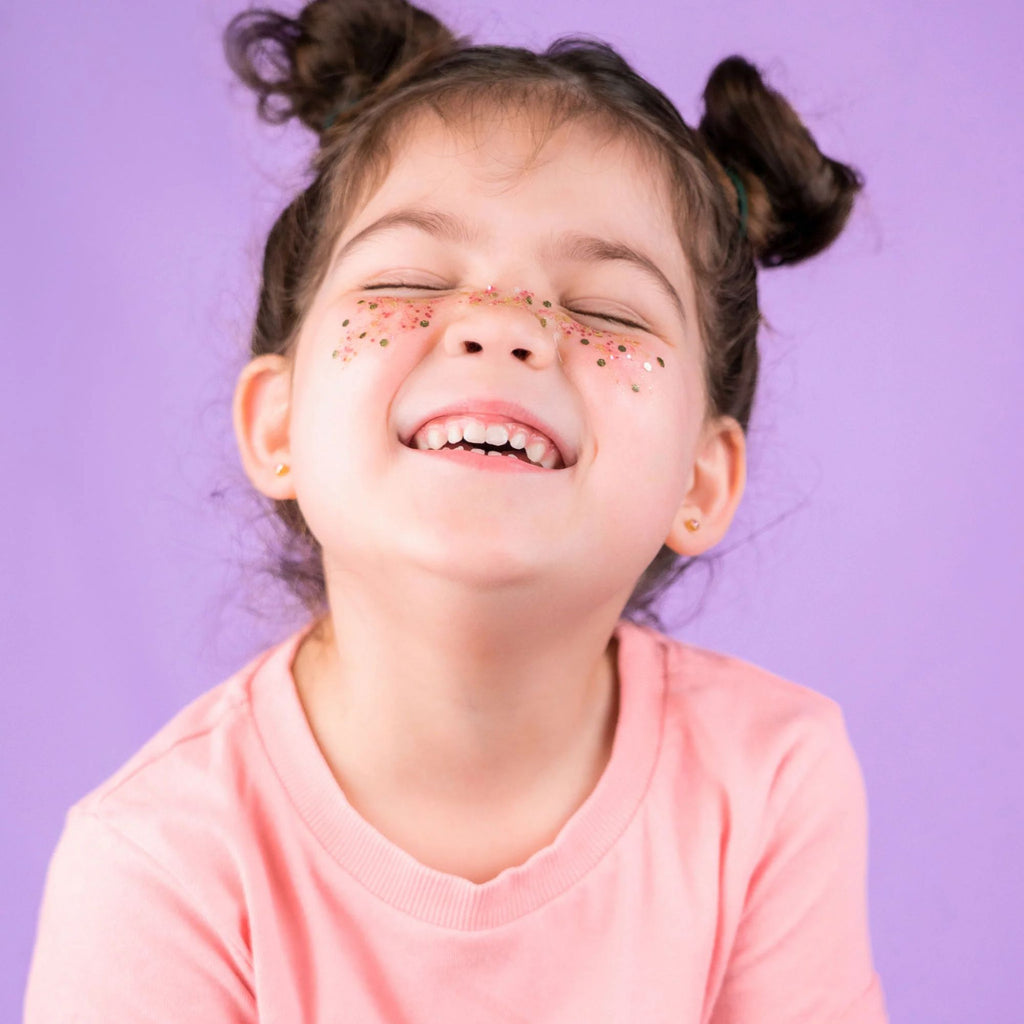 A girl with two space buns a pink top with glitter on her cheeks smiling with her eyes closed against a purple background. 