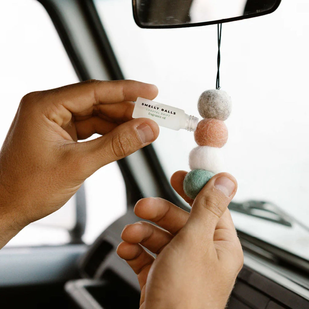 Hands in a car refilling a felt ball car air freshener hanging from a rearview mirror with fragrance oil