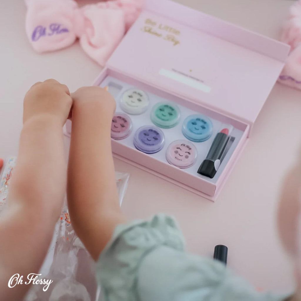 Two kids hands reaching to a Pink box of kids makeup set with 6 shades of eyeshadow and a lipstick with pink headbands on the table