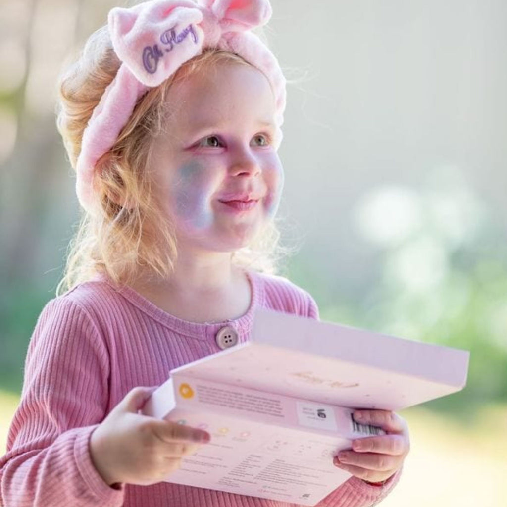 Little girl smiling wearing a headband  with eyeshadow on her face holding a pink makeup set