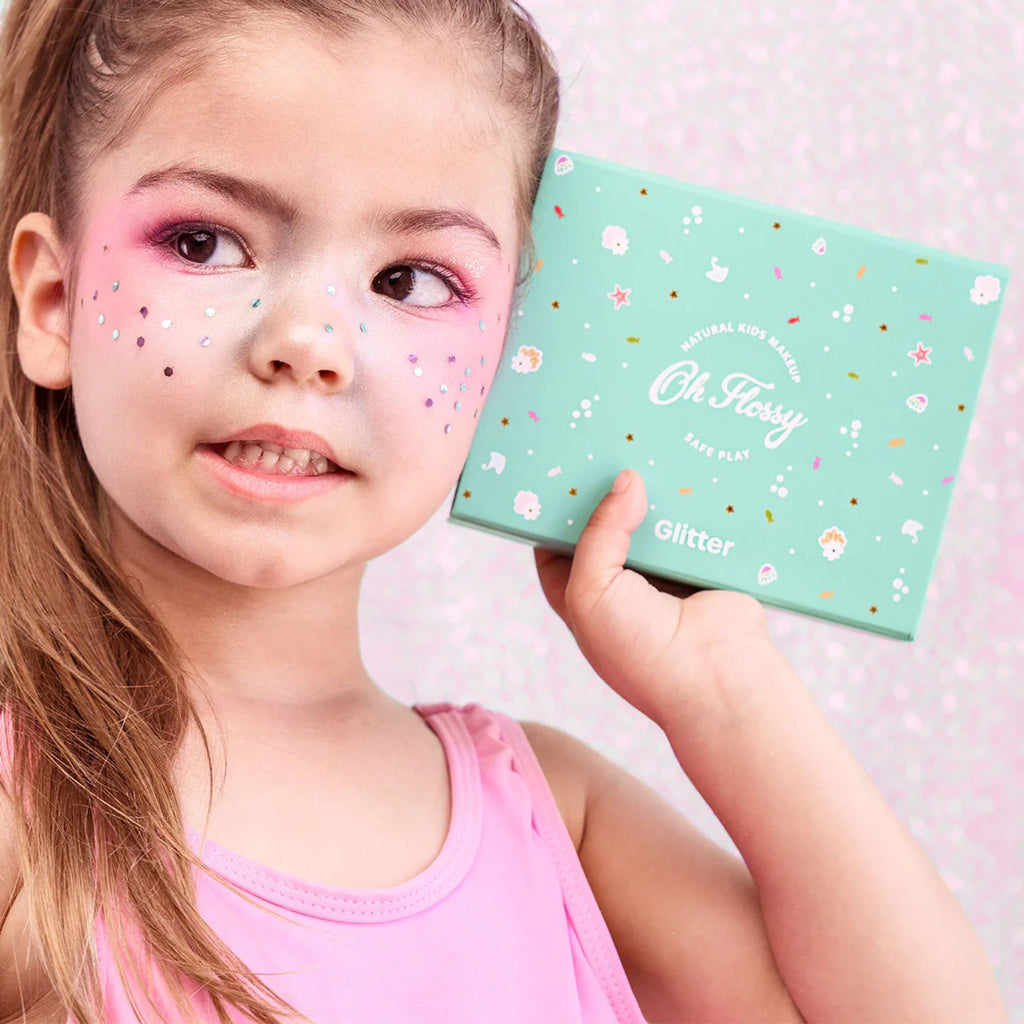 A young girl with. pink eyeshadow and face sparkles on her cheeks holding a blue box  against her face labeled "natural kids makeup safe play, oh flossy, glitter"