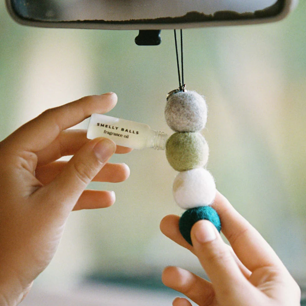 Hands in a car refilling a felt ball car air freshener hanging from a rearview mirror with fragrance oil