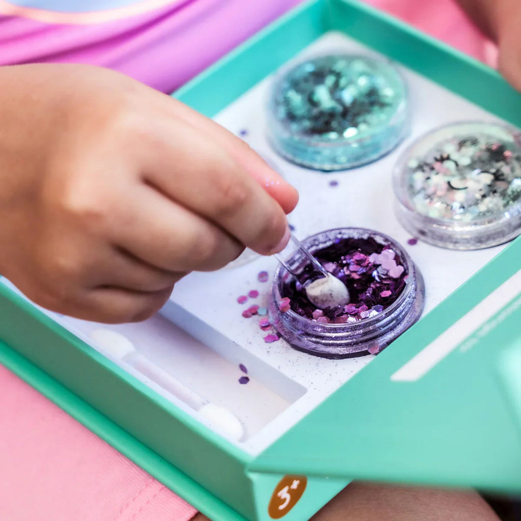 A blue box with three pots of purple, blue and white glitter with a child's hand using an applicator in the purple glitter