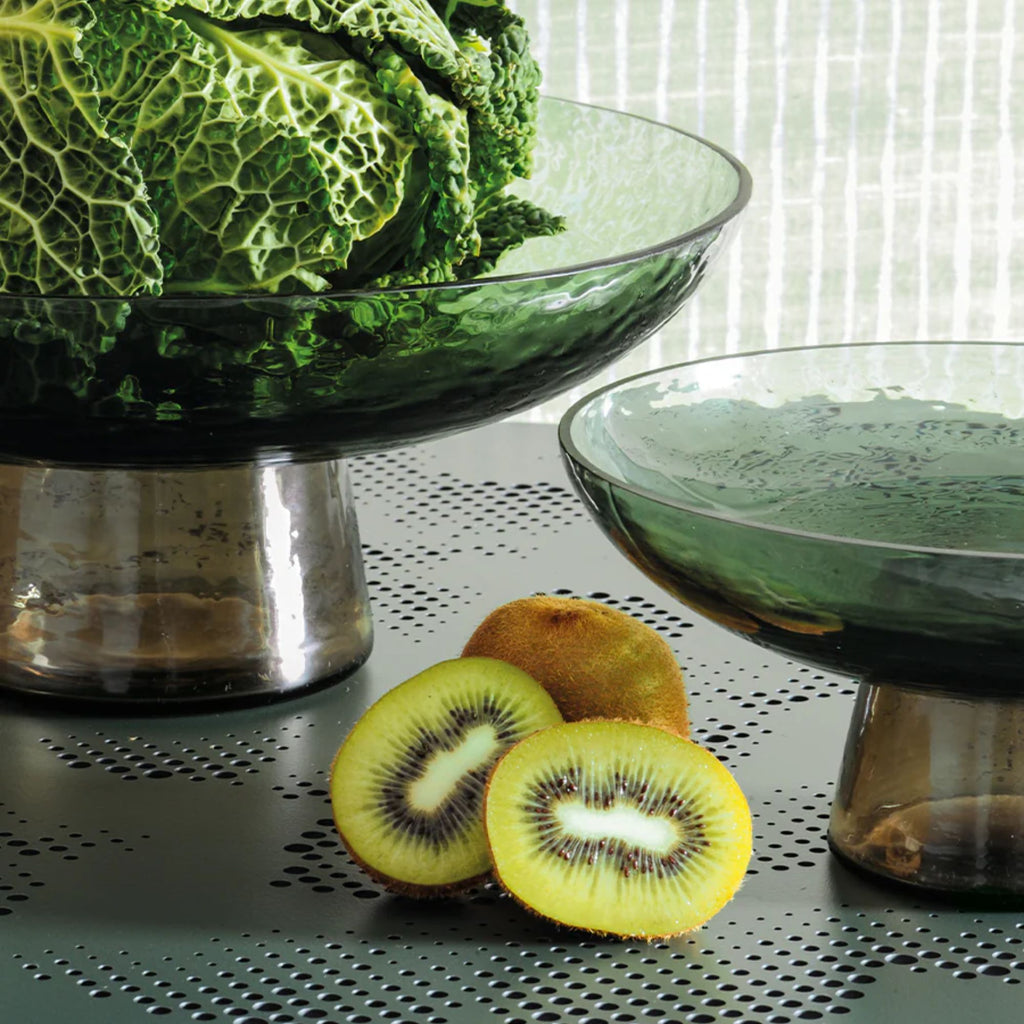 A large and small hammered texture green glass on a bowl on a brown glass stand against a textured background on a table, with cabbage in the large stand and a stack of sliced kiwifruit on the table.