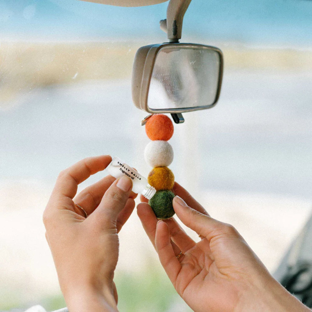 Hands in a car refilling a felt ball car air freshener hanging from a rearview mirror with fragrance oil