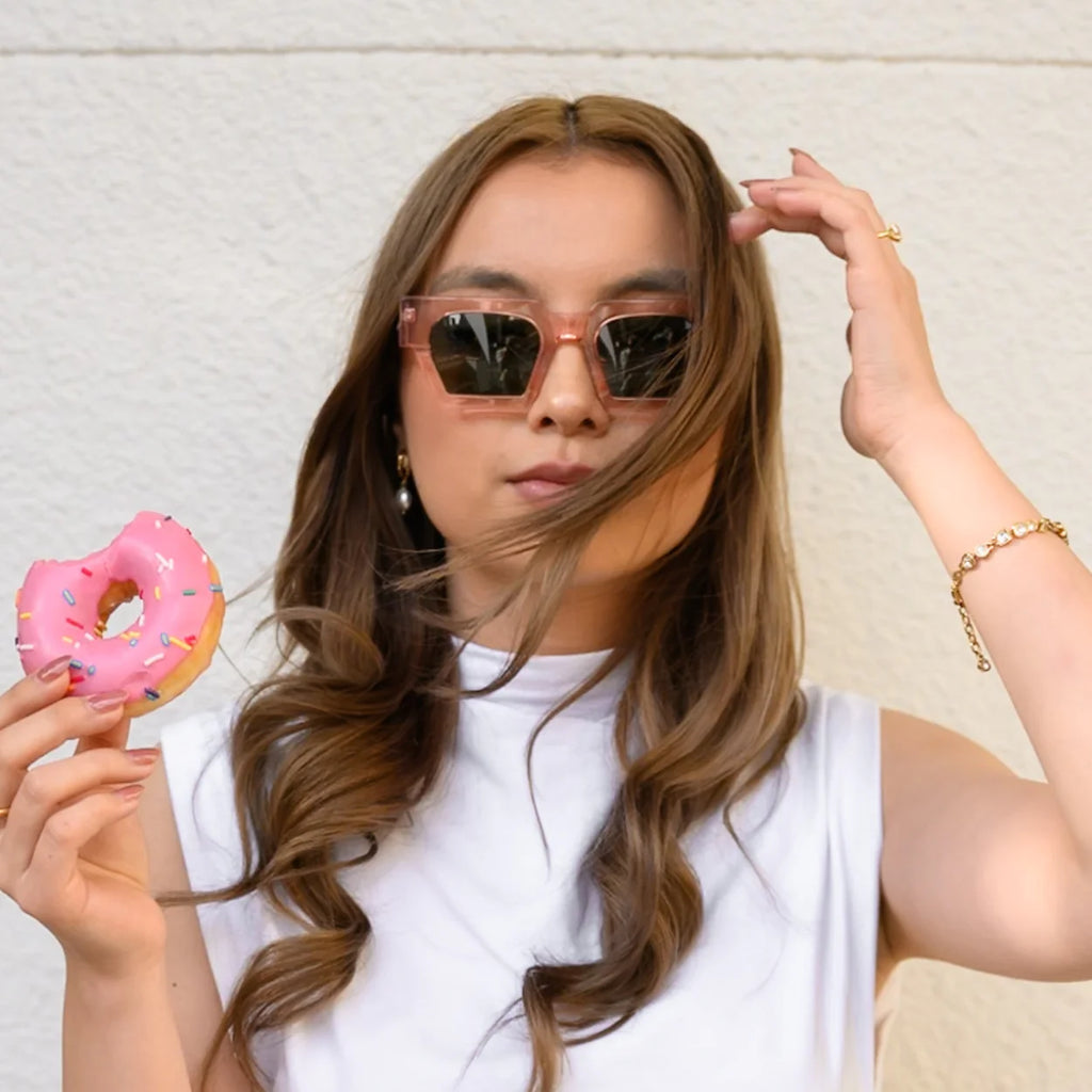 A woman wearing sunglasses holds a pink donut and touches her hair 